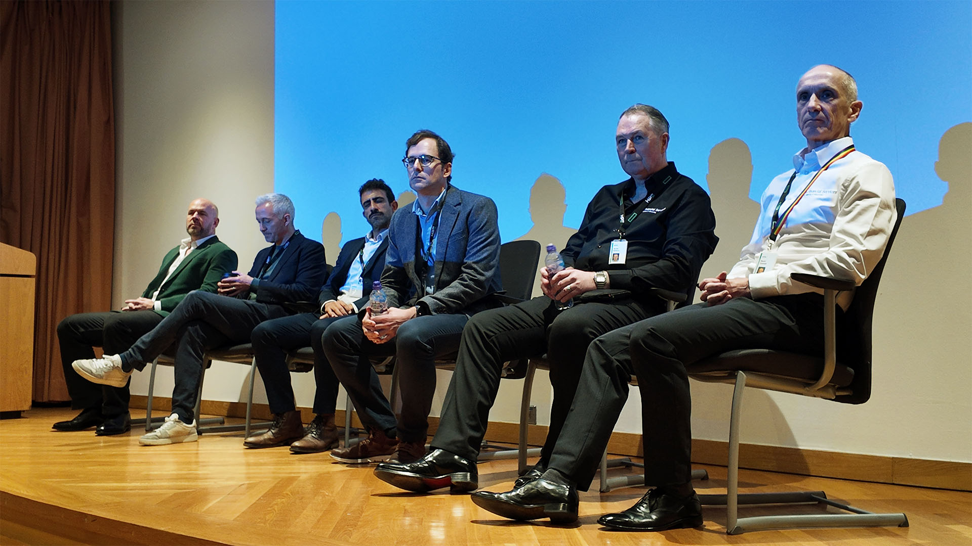 Six men sit in chairs on a stage for a panel discussion with a blue screen in the background