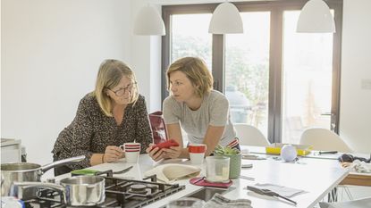 An older woman and her adult daughter look at a smartphone together in a kitchen.