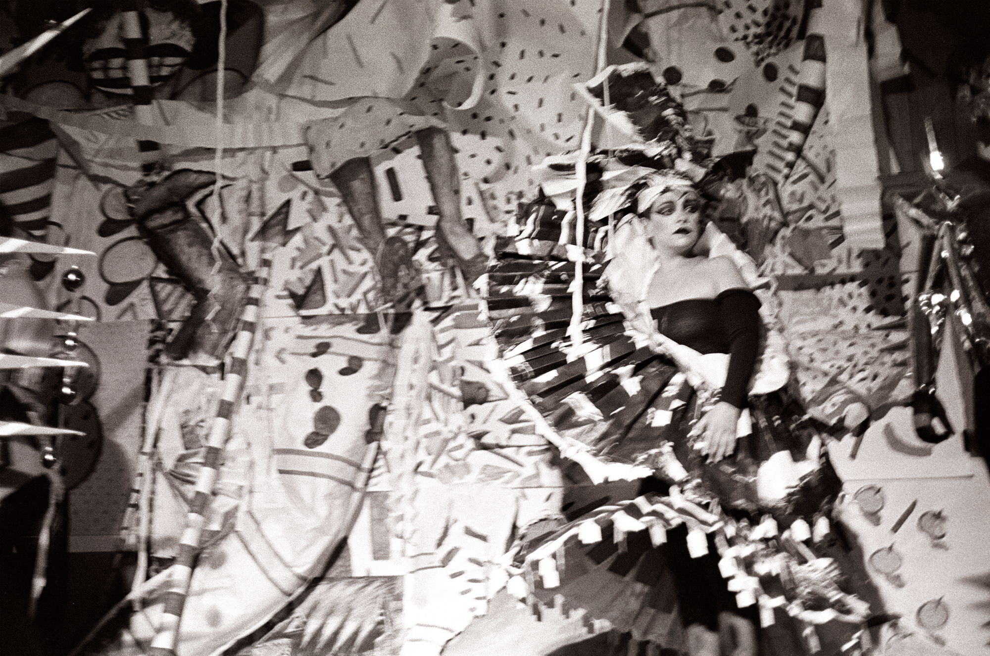Black and white photograph of a performer in elaborate paper costume and headdress surrounded by a chaotic backdrop of patterned paper and props.