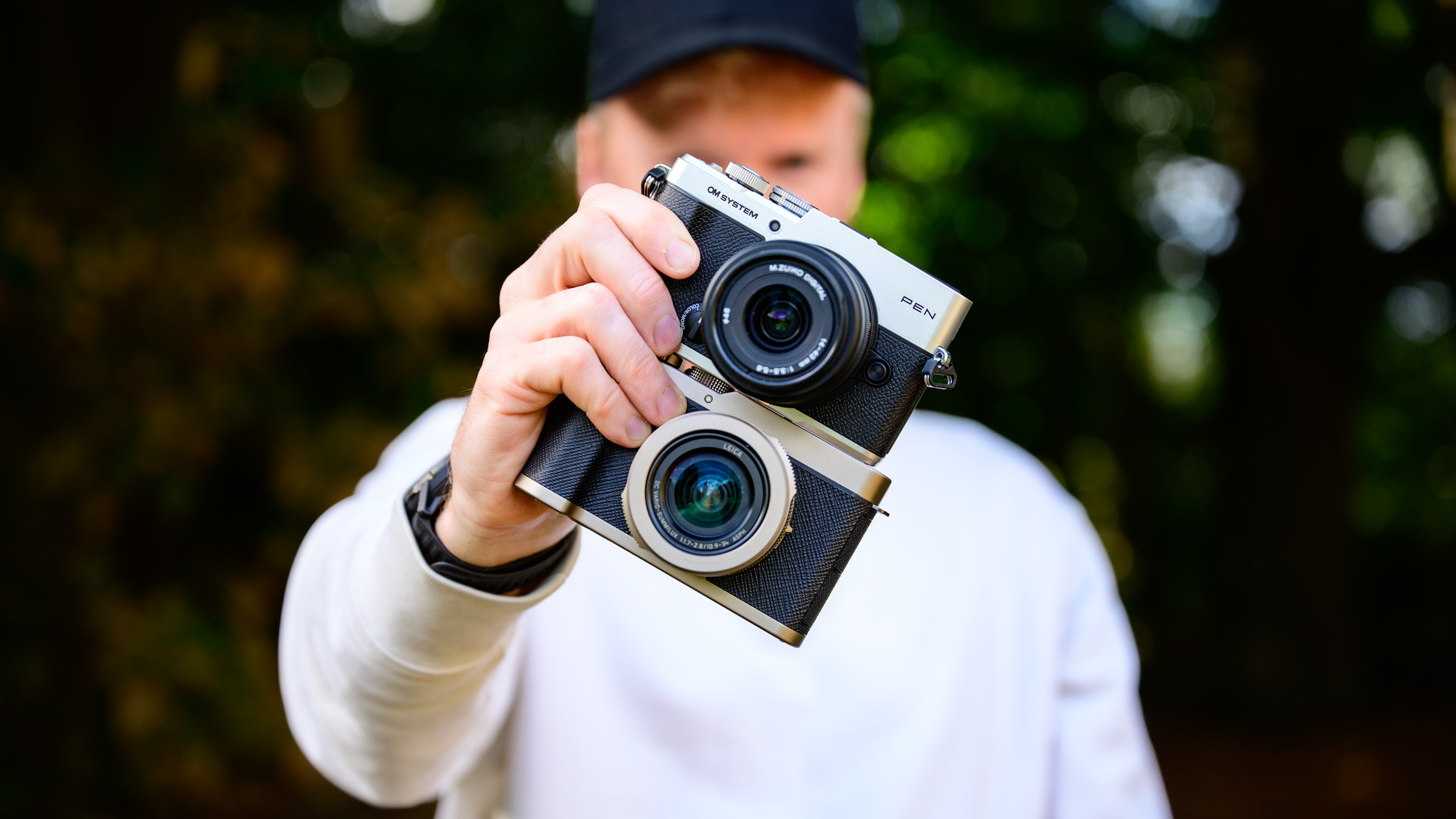 OM System PEN vs Panasonic Lumix L10: two micro four thirds cameras side by side in tech journalist Tim Coleman's hands, dappled tree light background
