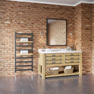 bathroom with exposed brick wall, black heated towel rail with wooden shelving, white basin with wooden cabinet underneath and large black rimmed mirror above