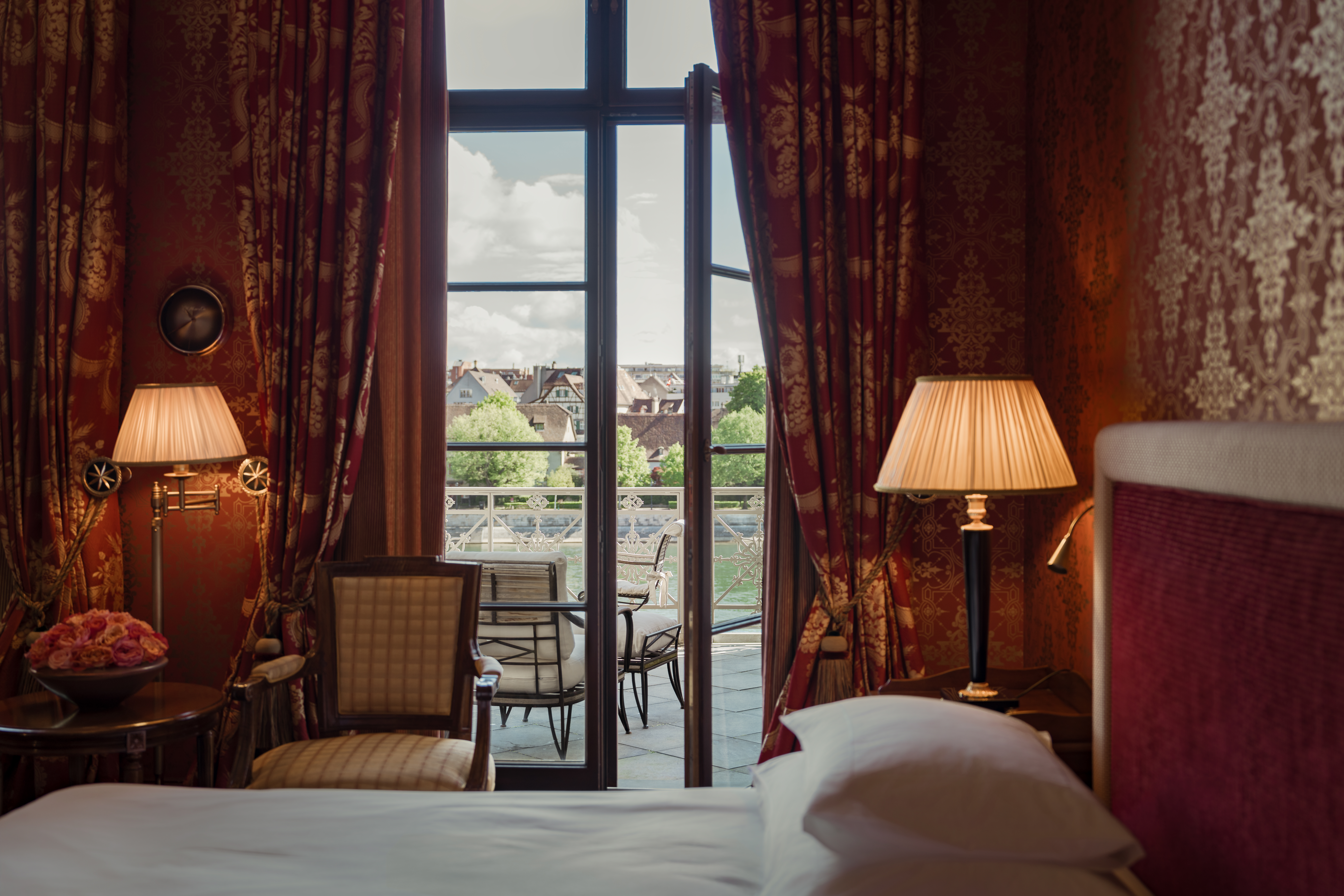 A room inside Les Trois Rois, with red wallpaper, old-fashioned lamps, and a white bedspread, with a balcony overlooking the Rhine.