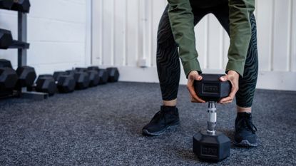 Woman lifting dumbbell from the floor by end, going into goblet squat, wearing leggings and workout shoes, doing cardio before or after weights