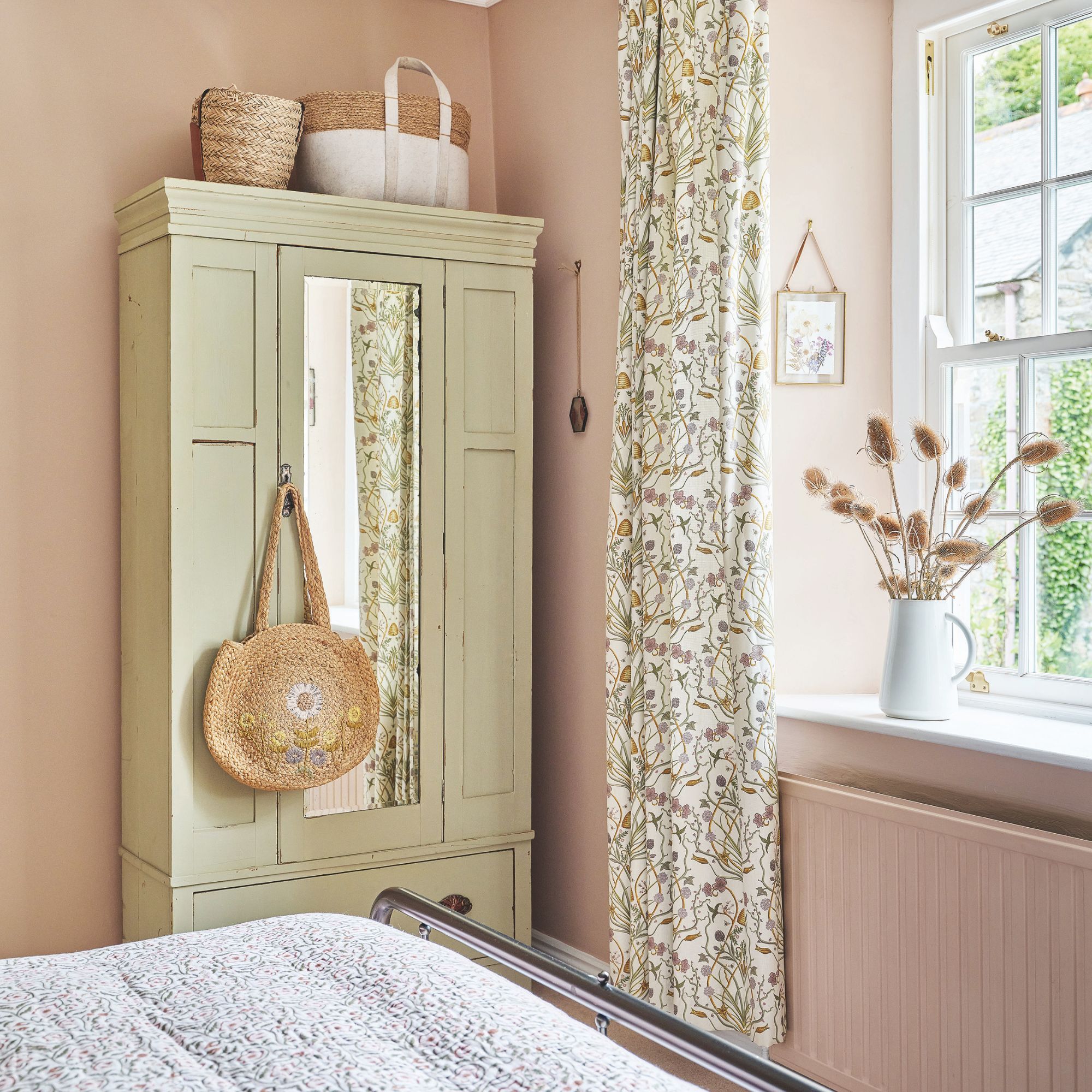 Pink bedroom with floral curtains around the window and a cream wardrobe with a wicker bag hanging from the door handle