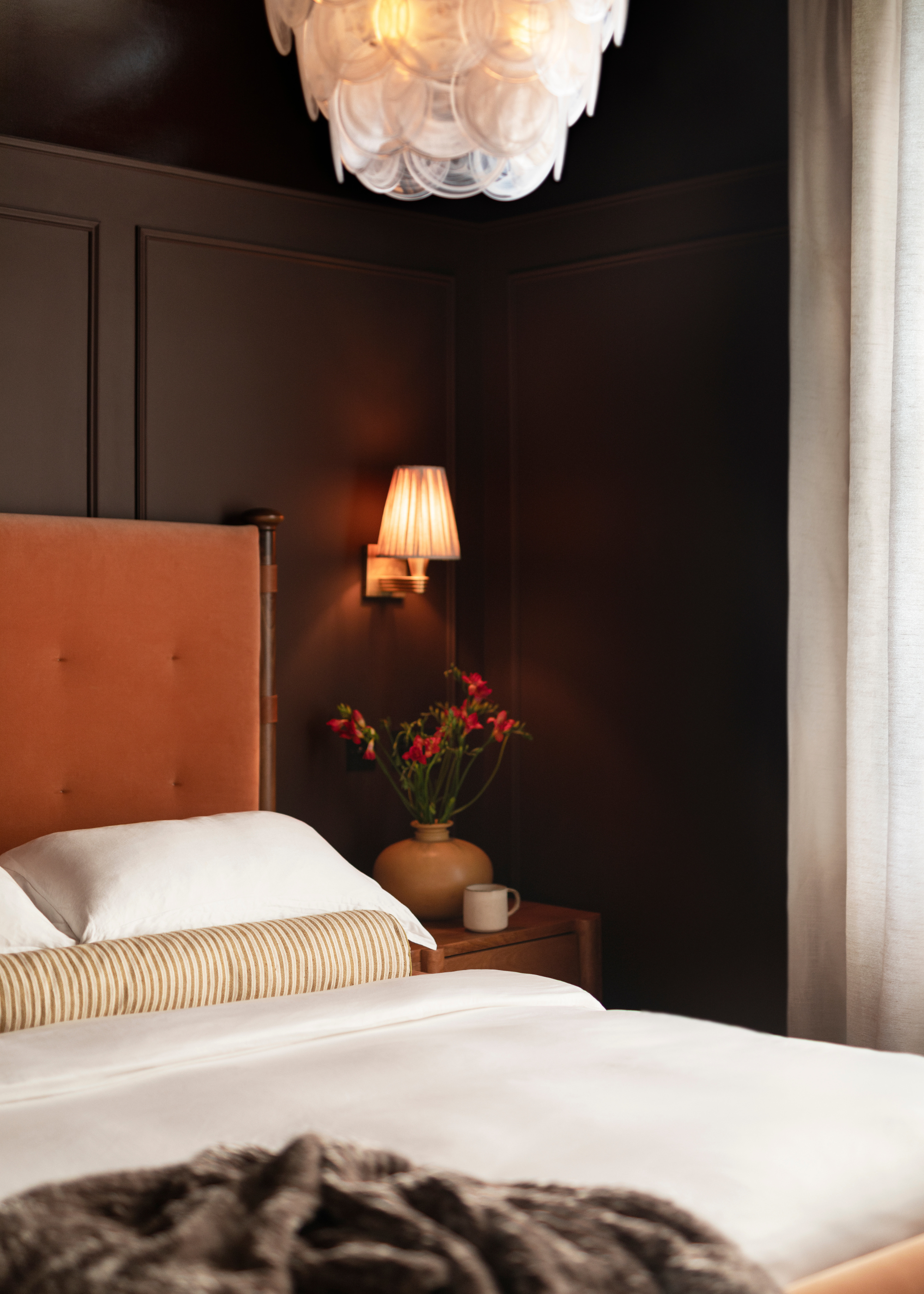 A dark brown bedroom with a murano-style chandelier, a bed with white bedding, a striped bolster pillow, a fuzzy blanket, and a vase of flowers by a ceramic vase on a nightstand below a wall lamp