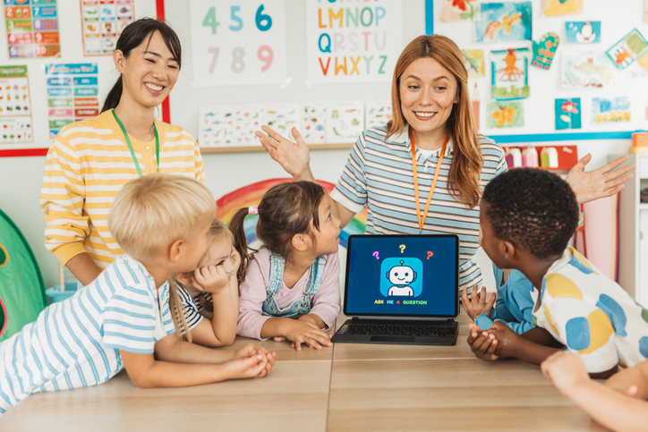 Female teacher is surrounded by her kindergarten students as she shows them how to use an ai on digital tablet.