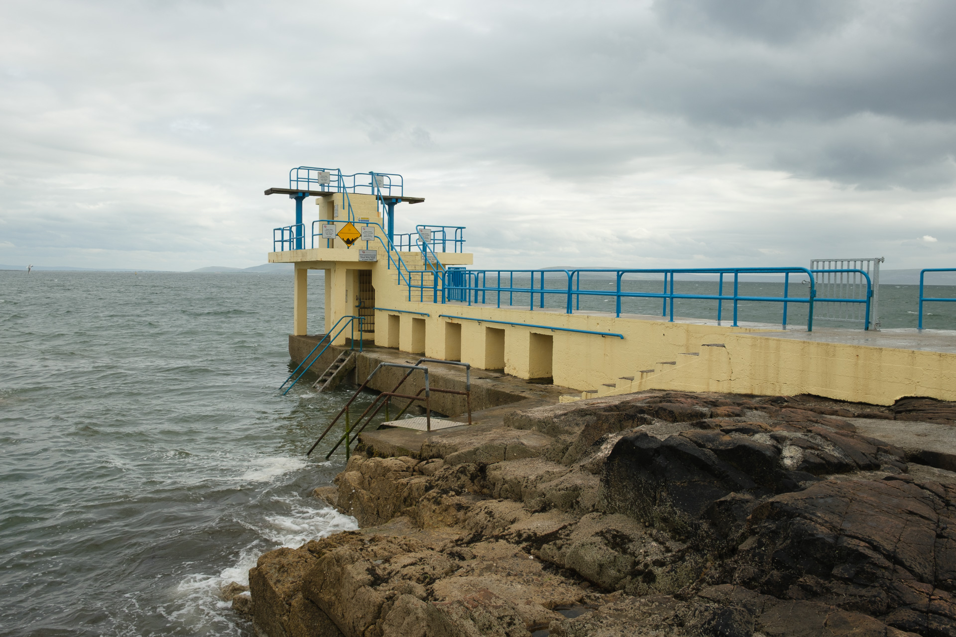 A cream diving tower with blue railings extends over a choppy sea beneath an overcast sky.