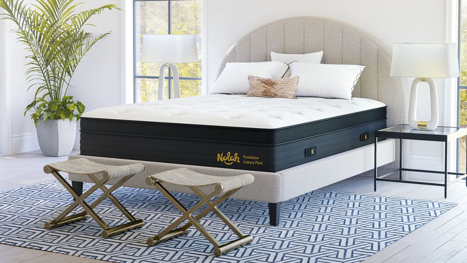 Nolah evolution mattress pictured in a white bedroom with light grey flooring, a white and blue geometric patterned rug, a large houseplant, two matching white lamps, and two large windows.