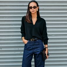 New York Fashion Week SS26 street style photo of a woman standing in front of a metal wall wearing a black headband, oval sunglasses, chunky earrings, a black v-neck sweater, black croc belt, and dark-wash jeans