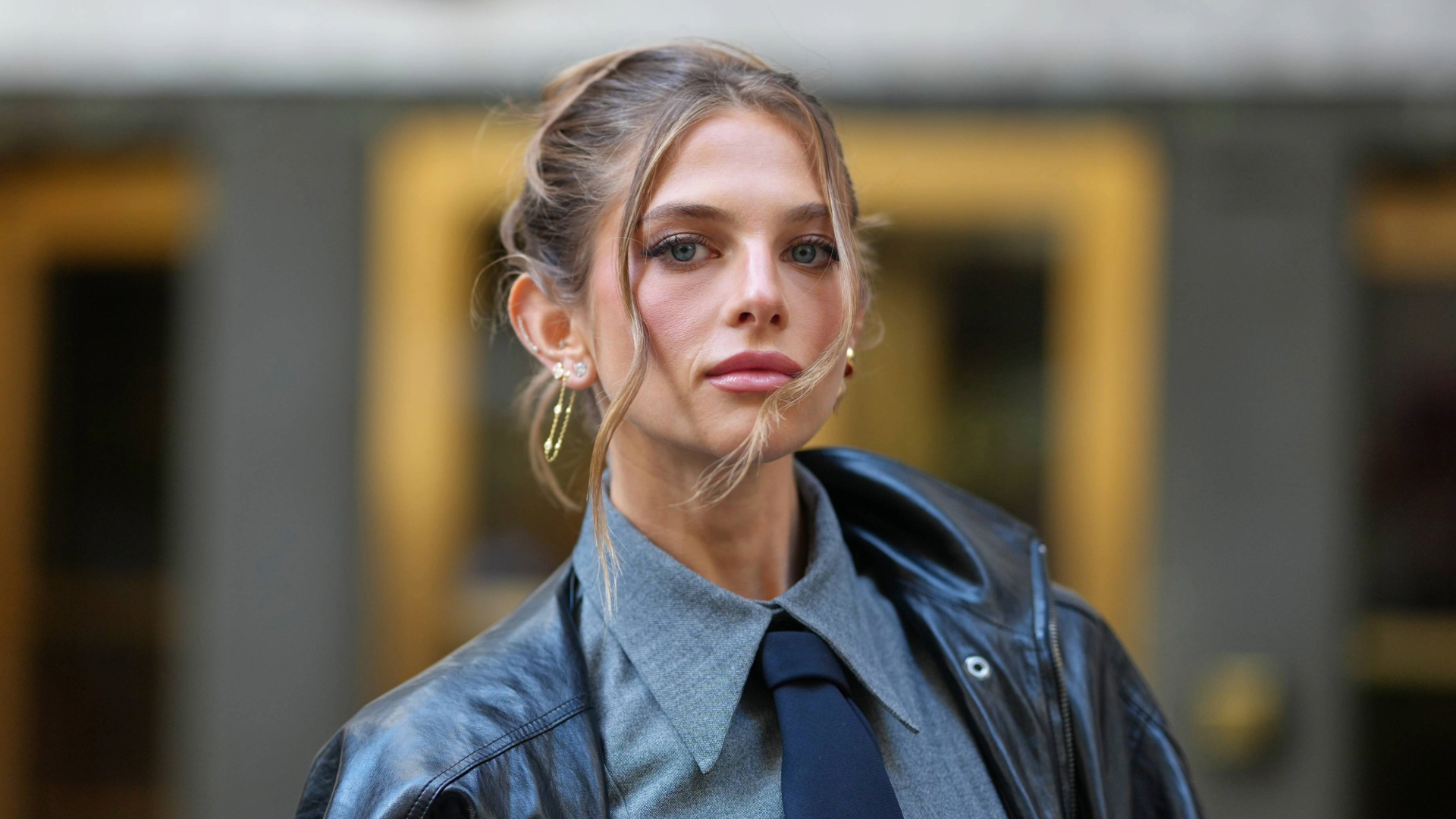 NEW YORK, NEW YORK - SEPTEMBER 15: A guest wears brown hair pulled back into a loose topknot with face-framing tendrils, small gold hoop earrings and multiple gold rings, an oversized black leather jacket, a grey button-down shirt with a pointed collar and a navy tie, outside TWP, during New York Fashion Week, on September 15, 2025 in New York, New York (Photo by Edward Berthelot/Getty Images)