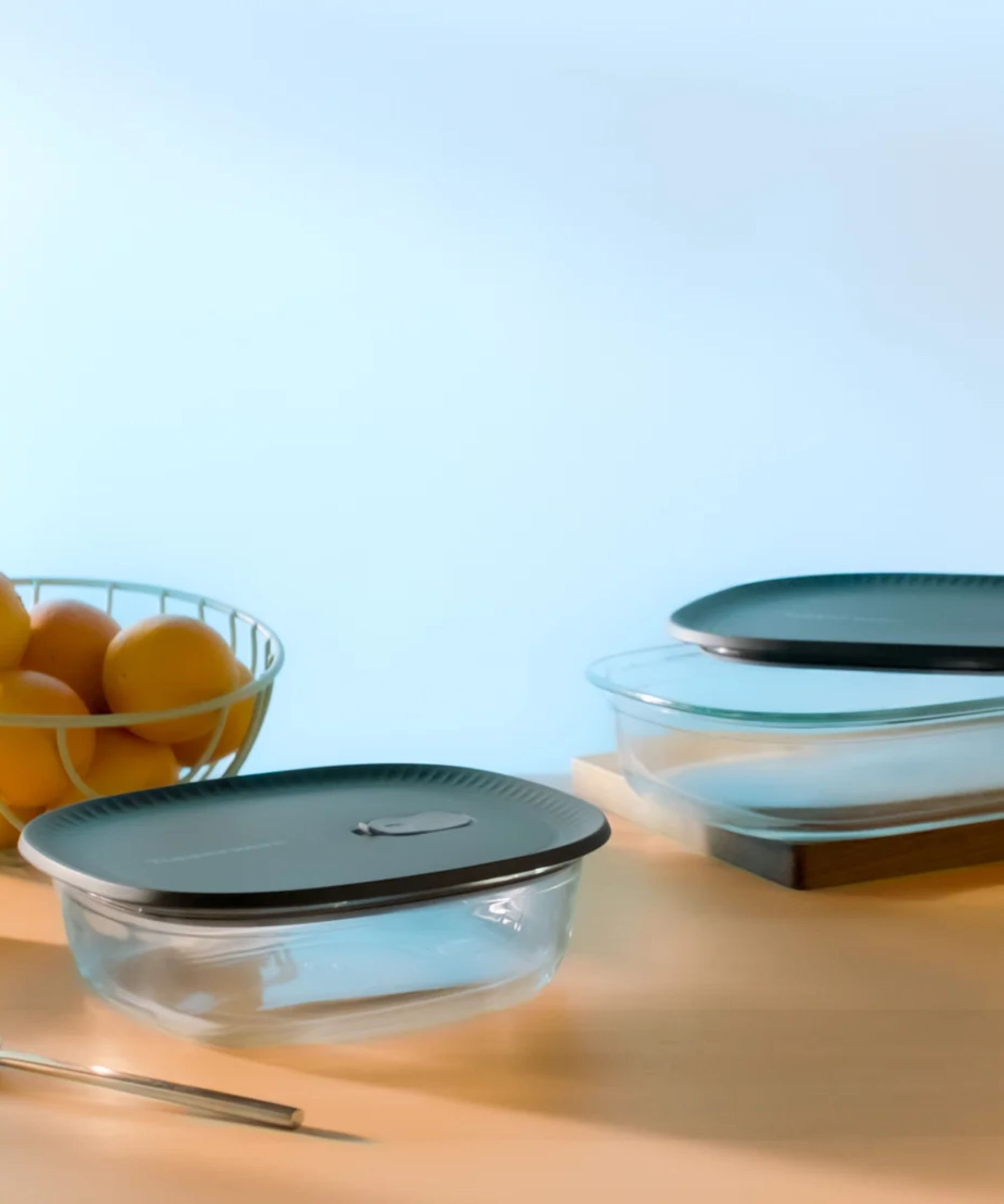 Glass food containers with navy lids on a wooden table, next to a wire fruit bowl full of oranges