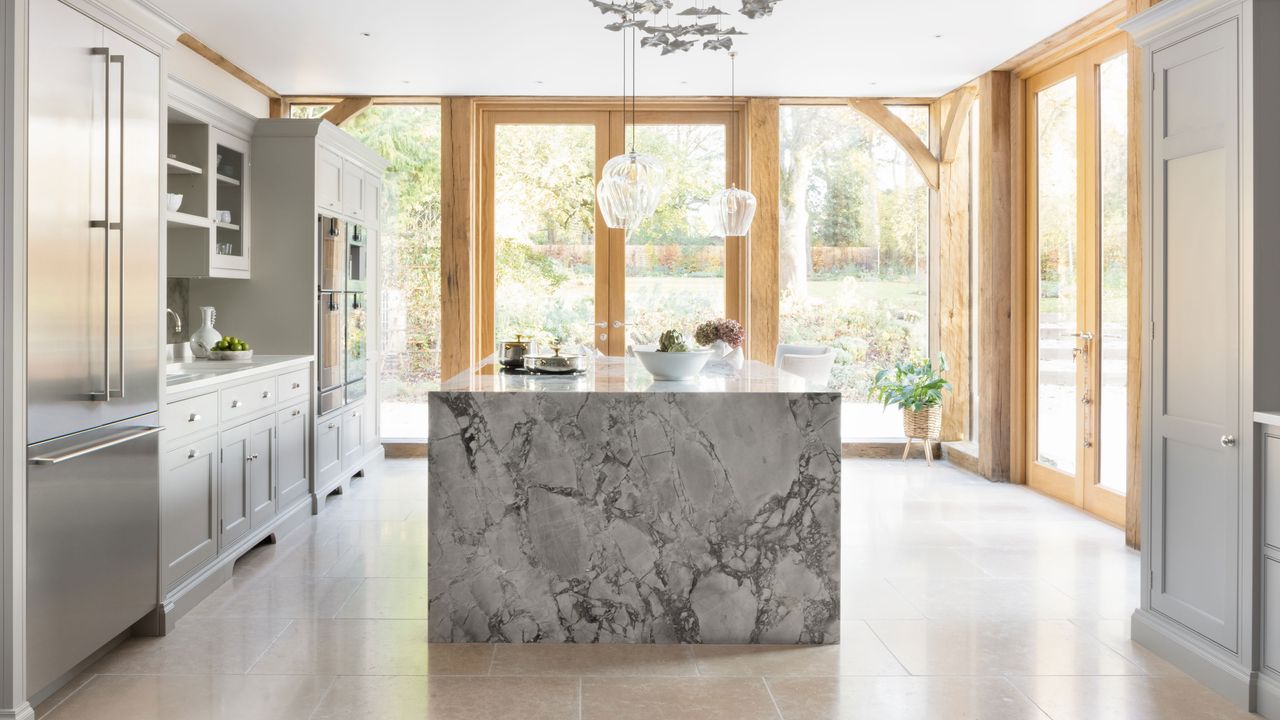 Island countertop ideas. Large white kitchen space with statement marble island, wooden joinery and door frames, large stone floor tiles, white kitchen cabinets, decorative hanging feature and lights over island