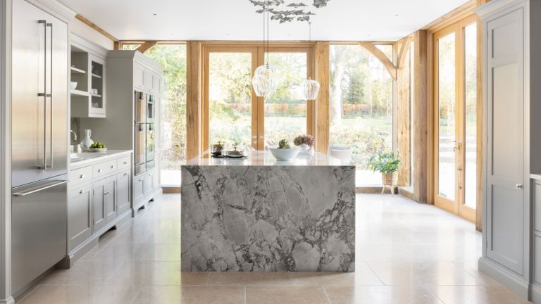 Island countertop ideas. Large white kitchen space with statement marble island, wooden joinery and door frames, large stone floor tiles, white kitchen cabinets, decorative hanging feature and lights over island