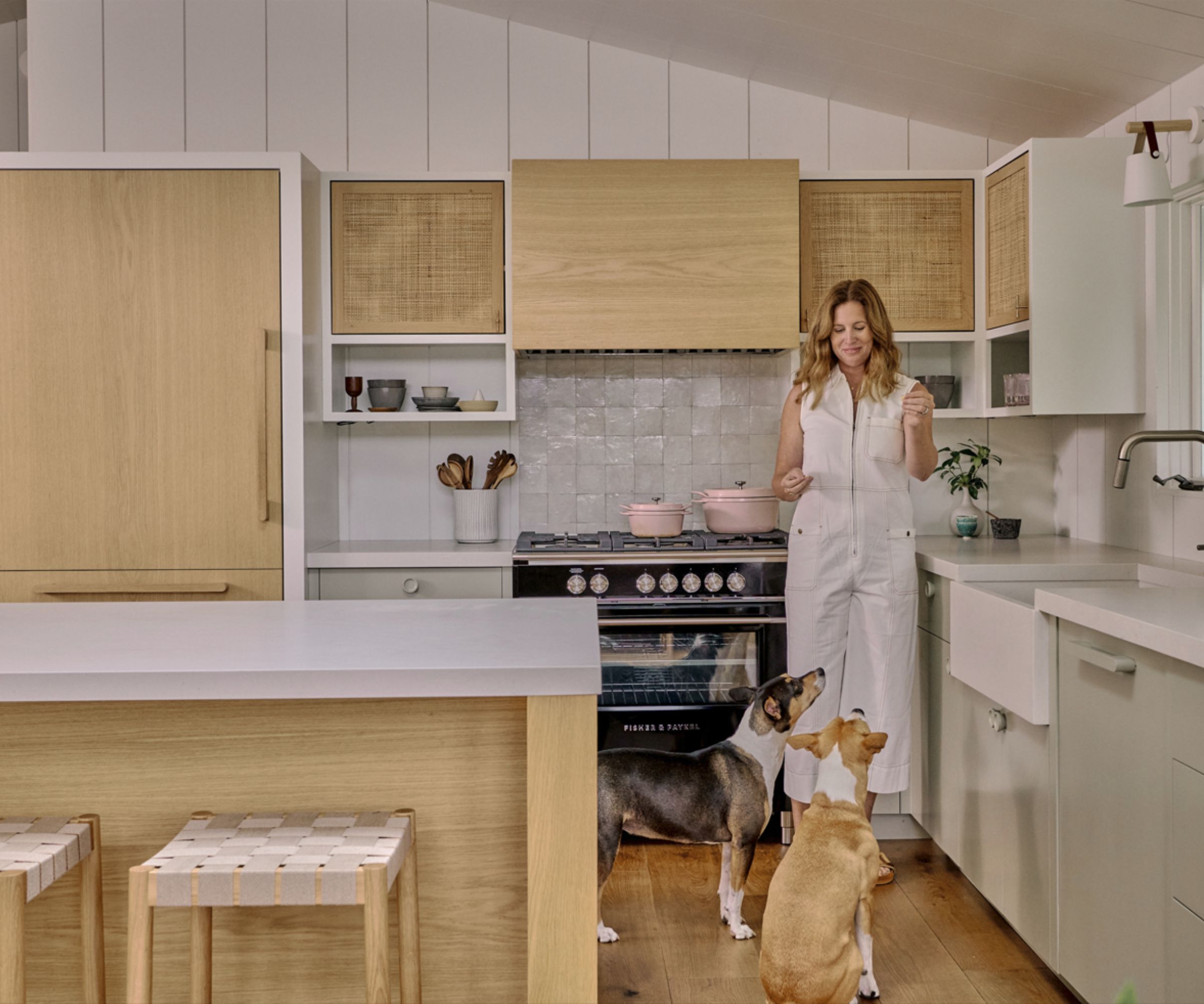 Jess Cooney standing in her lakehouse kitchen with two dogs, warm wood kitchen island and cabinetry, gray tiles, wooden flooring