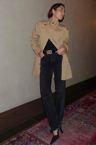 spring trench coats shown in a photo of a French woman standing in a hotel hallway wearing a cropped trench coat with a black blouse, black belt, black jeans, and black pointed pumps