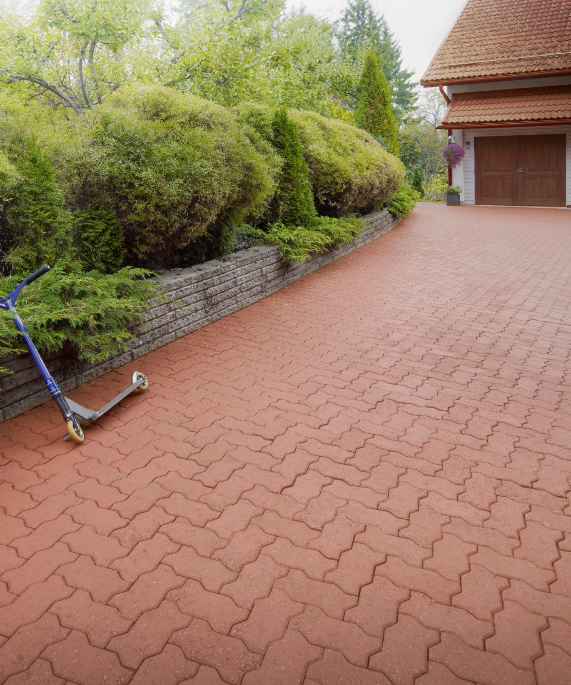 Herringbone-patterned brick driveway beside a low retaining wall, conifers, and a scooter leaning against it.