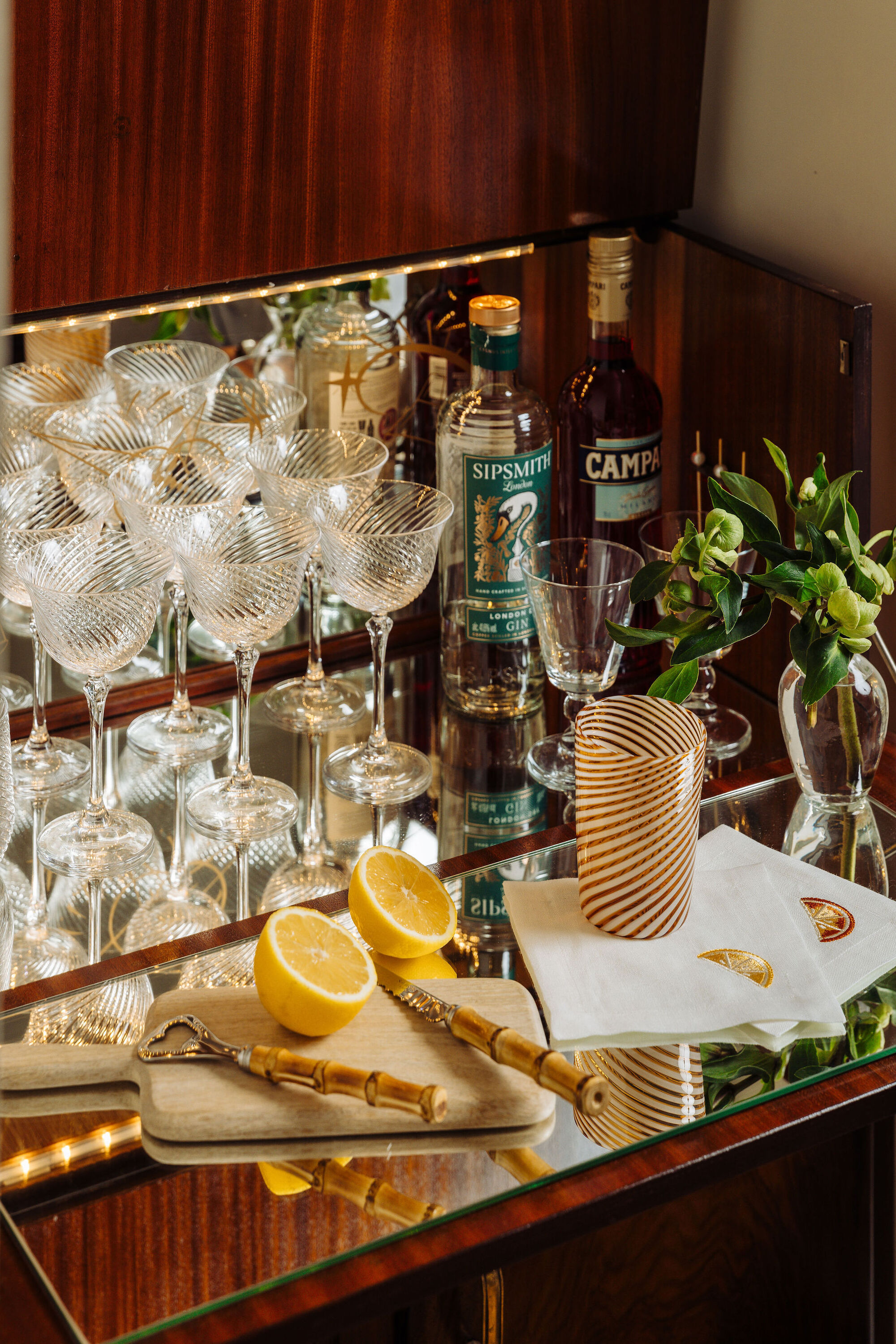 close up of home bar with mirrored backsplash, bamboo bar accessories, striped venetian glass, spirits, a vase with branch, and glassware