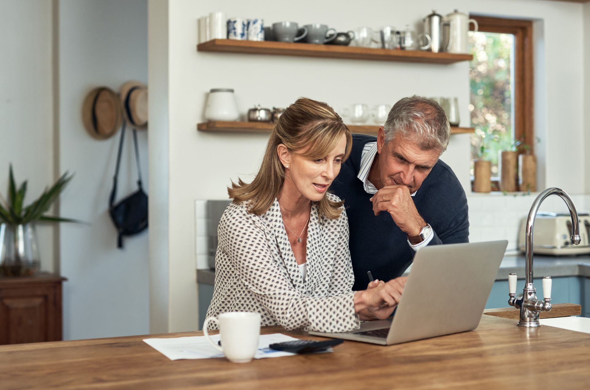 A senior couple sits at the kitchen table in a modern home working on a laptop.