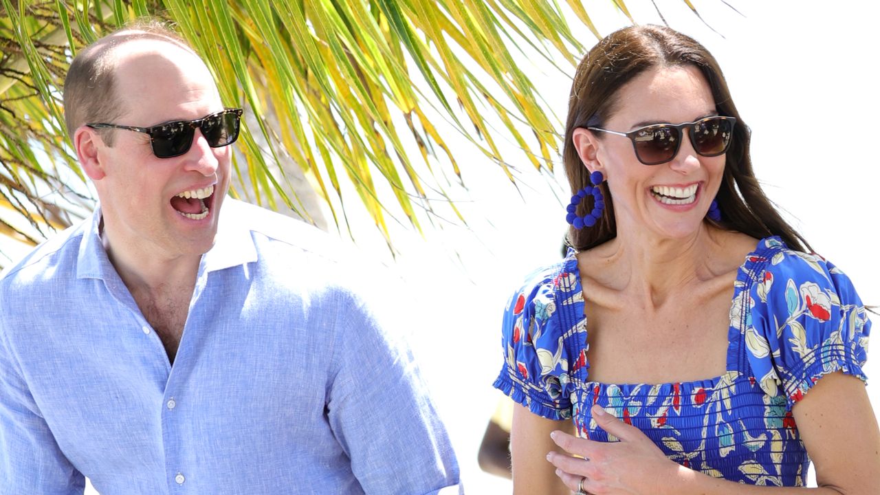 Prince William and Kate Middleton on a beach in front of a palm tree, they are both wearing sunglasses, William wears a light blue shirt and Kate wears a dark blue dress with floral pattern