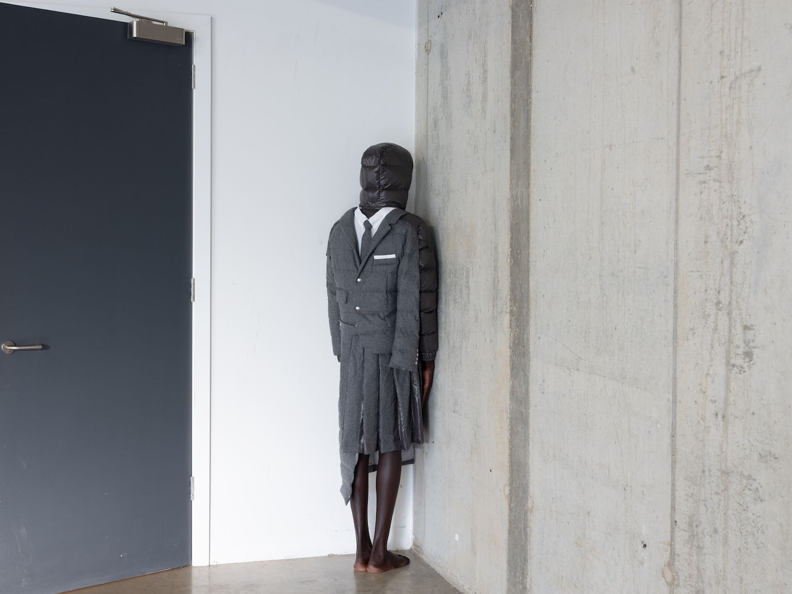 Man faces into corner of white wall in jacket which has suit jacket, tie and shirt on the back