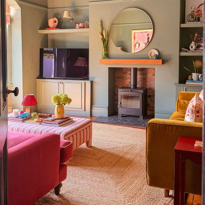 Sage green living room with jute rug, striped ottoman coffee table, wood burner and round wall mirror