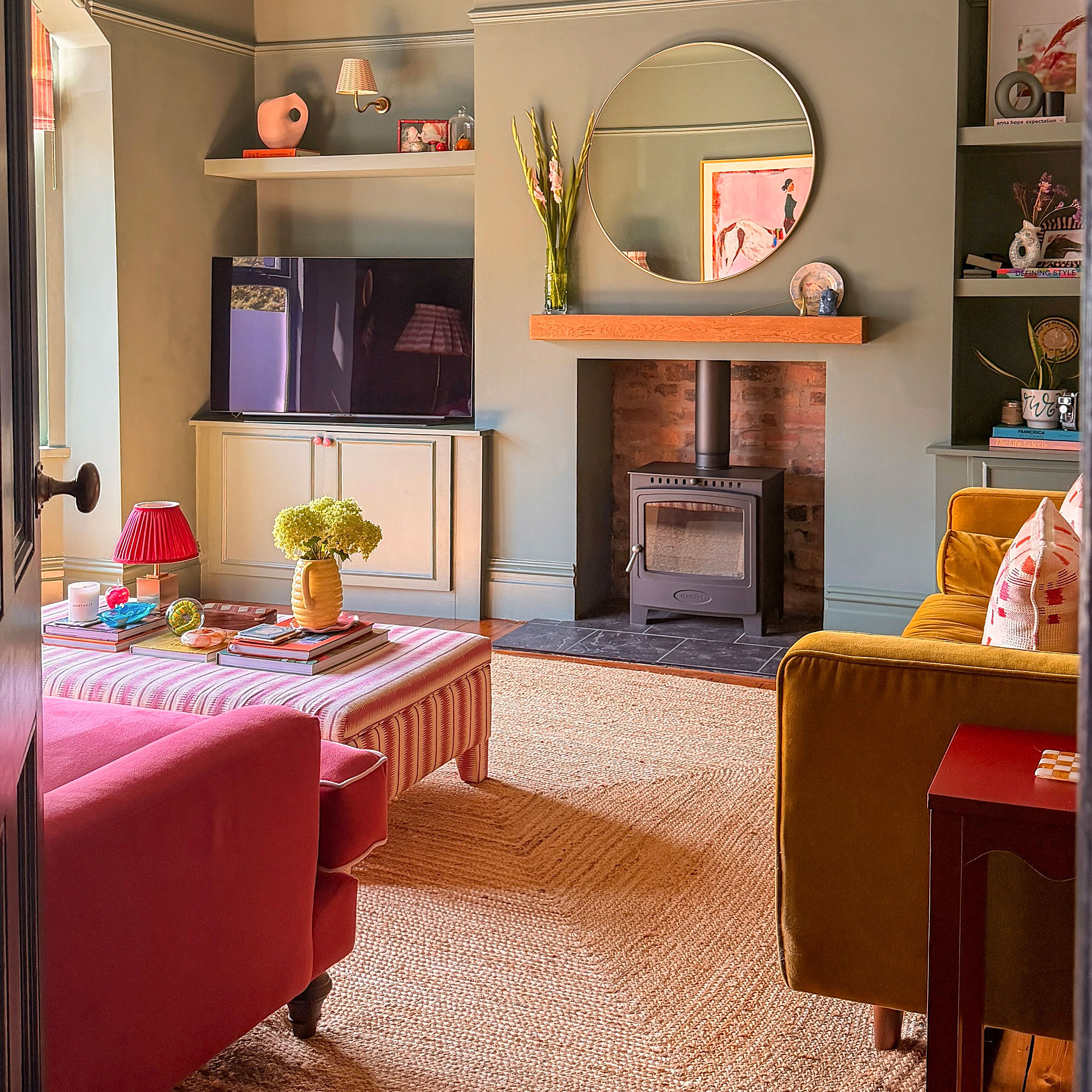 Sage green living room with jute rug, striped ottoman coffee table, wood burner and round wall mirror