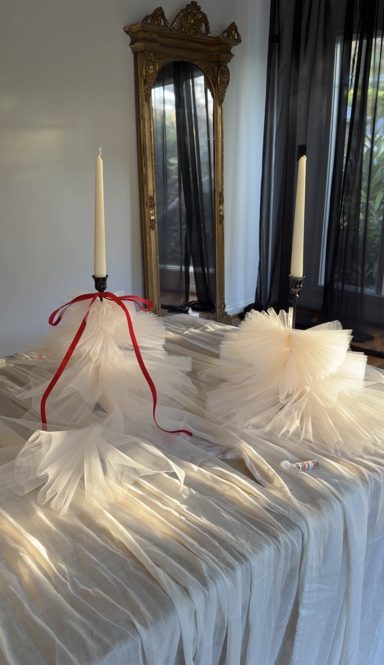 A dining table with a ruched white tablecloth and two black taper candle holders with candles with toile embellished skirts and one has a thin red velvet bow