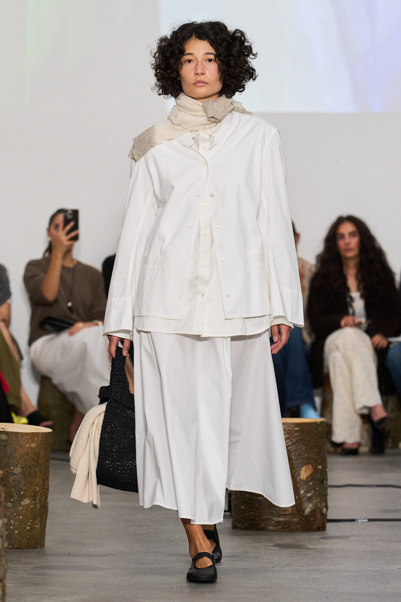 Layering staples for spring 2026 are shown in a photo from Aiayu's runway show, with a model walking while wearing an oversized white button-down shirt layered on top of another button-down styled with a white linen circle skirt, creme scarf, black ballet flats, and a black tote bag