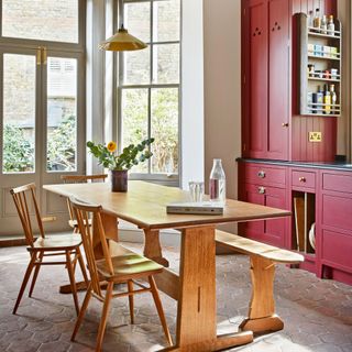 a kitchen with dark red cabinets with brass cup pulls, a wooden dining table and match ing spindleback chairs and a yellow pendant overhead