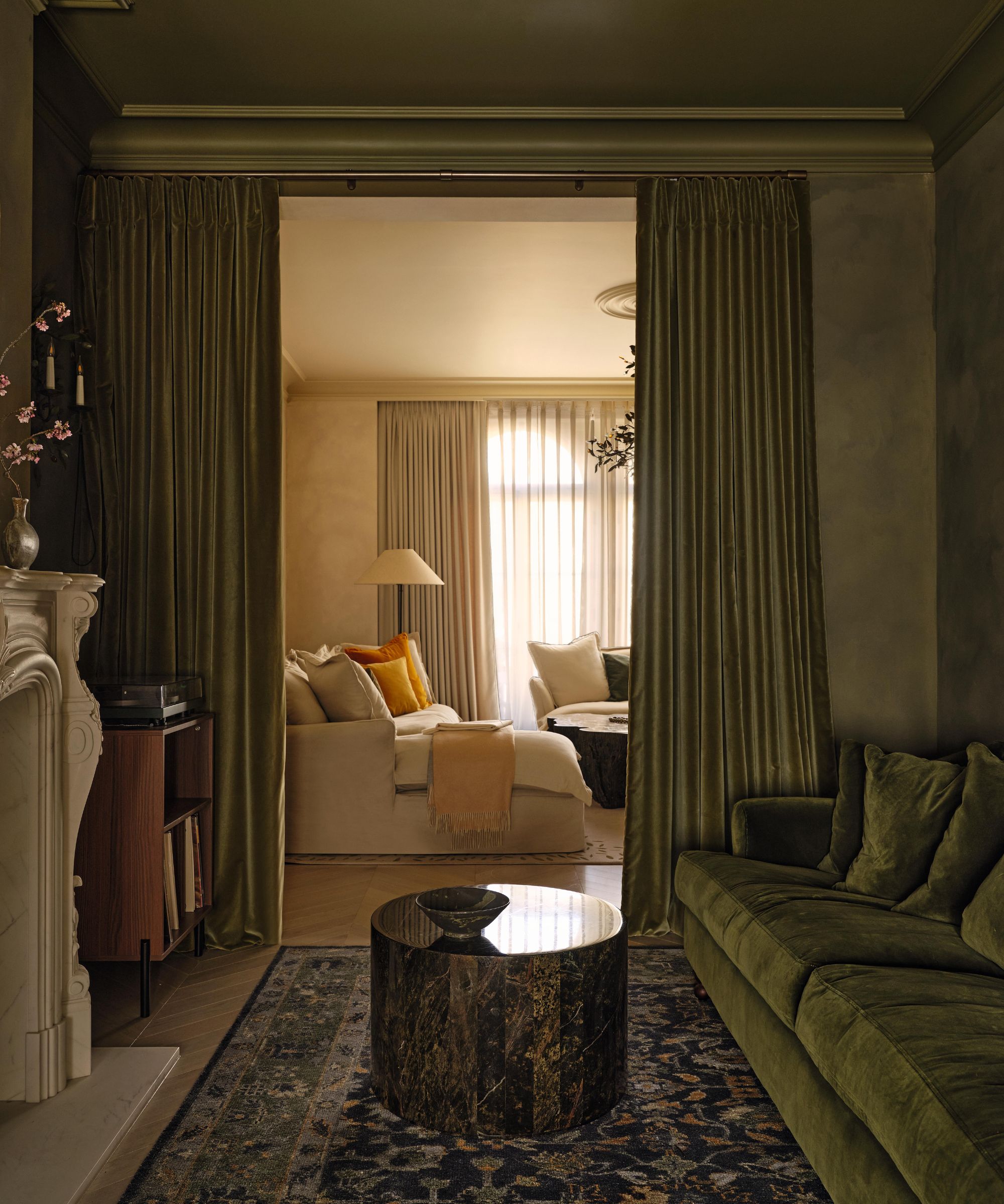 a limewashed green sitting room with velvet green couch, marble fireplace, antique rug and green drapes on an opening leading through to another living room
