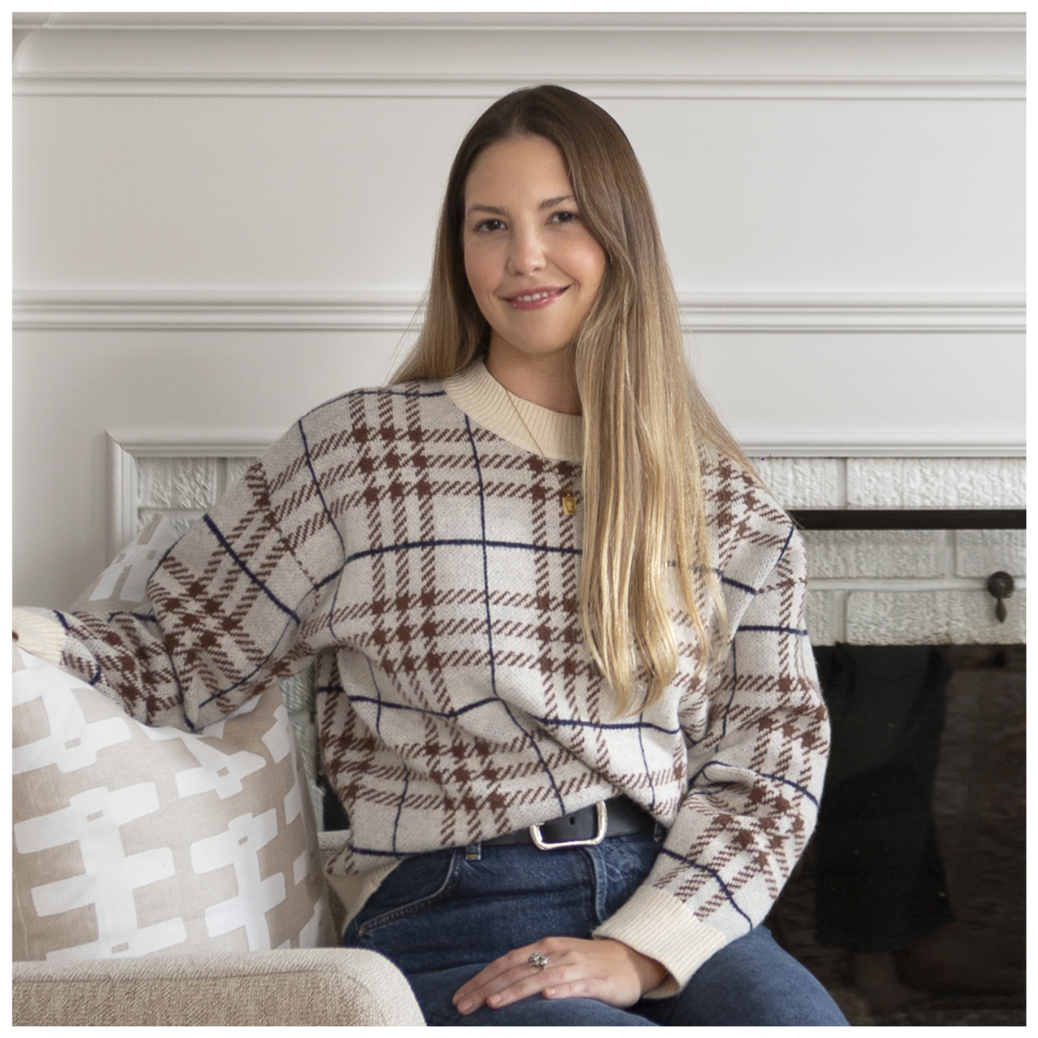 A headshot of a lady with ombre brown and blonde hair in a checked sweater and jeans, sitting on a beige sofa chair with a patterned pillow