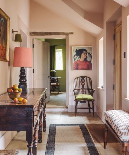 Hallway in a historic home color-drenched plaster pink with sloped ceilings. A large wooden console table with a pink lamp, stone flooring with a jute runner rug, vintage artwork and an antique chair.