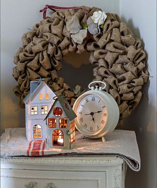 Two small christmas houses on a bedside table beside an alarm clock and brown wreath