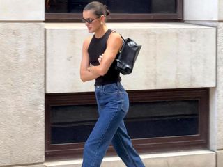 Female fashion influencer Valeria @sobalera walks the London sidewalks with a slicked back bun, oval sunglasses, statement earrings, a black racerback tank, a black shoulder bag and straight leg jeans