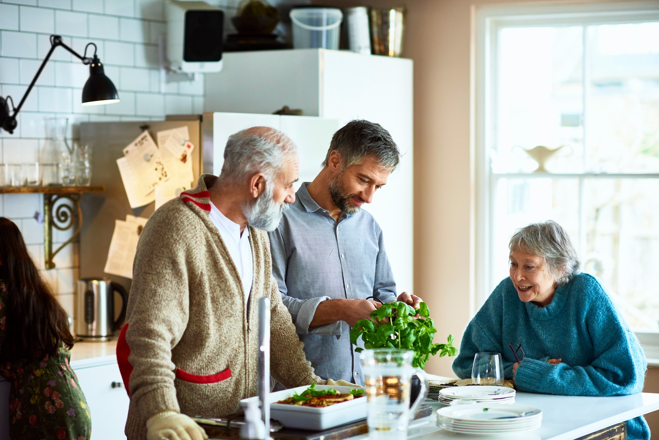 Mature man and woman visiting their son on the weekend, cutting fresh basil from the plant and garnishing home made meal, crockery on kitchen island.