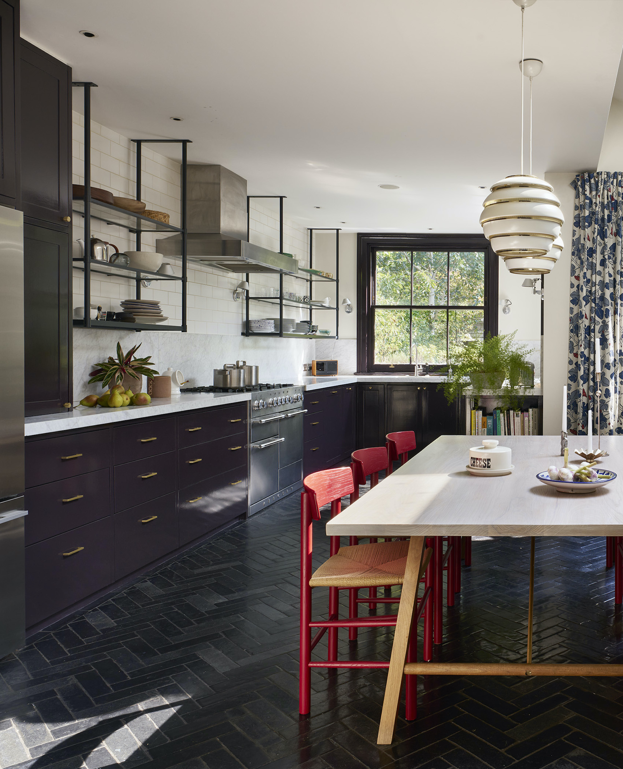 J-shaped kitchen with dark plum cabinetry, pale countertops and open metal shelving surrounding an ash dining table with red and natural wood chairs, above dark herringbone flooring.