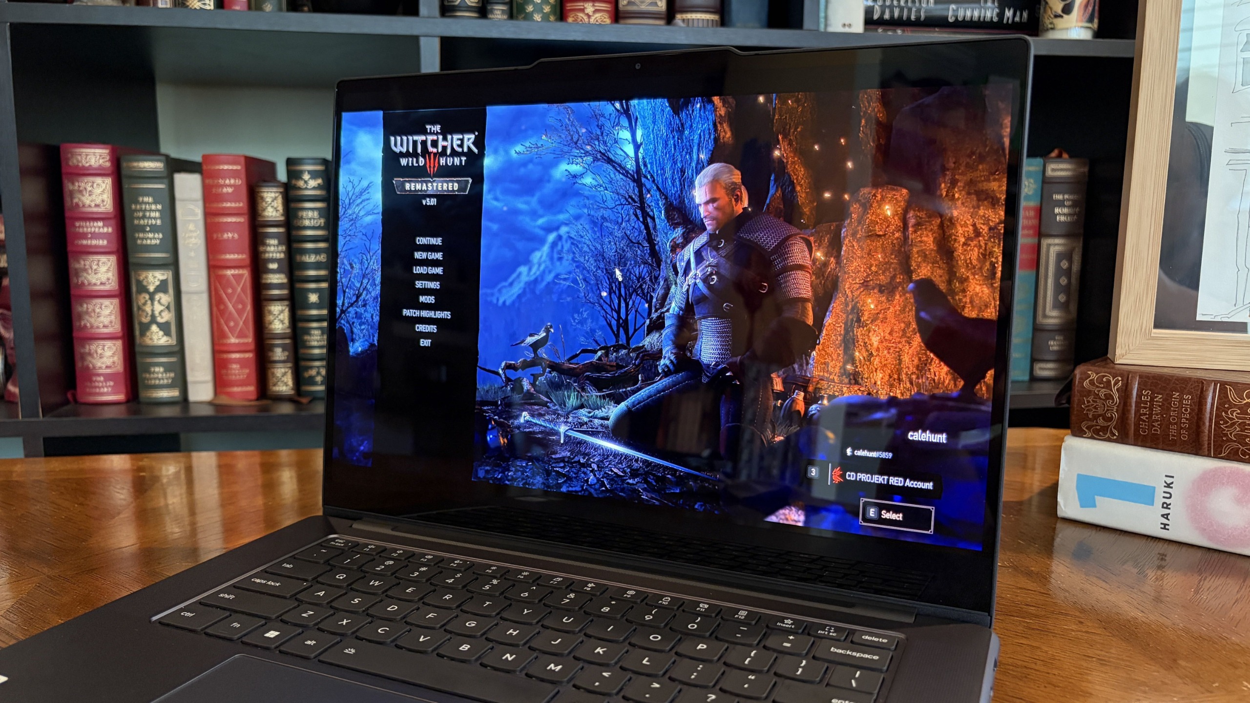 A Lenovo Pro 9n laptop on a wooden desk displays The Witcher 3 menu, with Geralt in a snowy blue forest beside a fiery orange scene; the dramatic contrast feels tense.