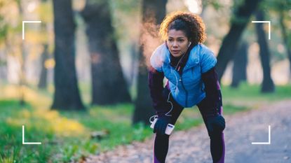 Woman bent over with hands on knees looking tired, wearing running clothes and headphones in the park, after wondering if she should work out with sore muscles