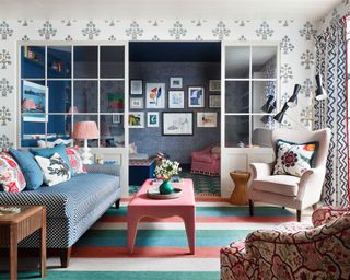 A broken plan living room with a colorful mixture of patterns in the choice of rugs, wallpaper, drapes and sofa fabrics.