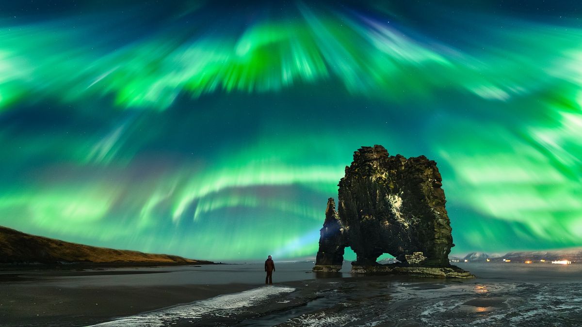 A person stands on a dark beach under a vibrant display of green and blue northern lights, which streak across the sky in a dynamic, swirling pattern. A large, rugged rock formation rises nearby, adding to the dramatic scene.