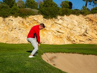 Alex Elliott playing a bunker shot from the top of the bunker with feet outside