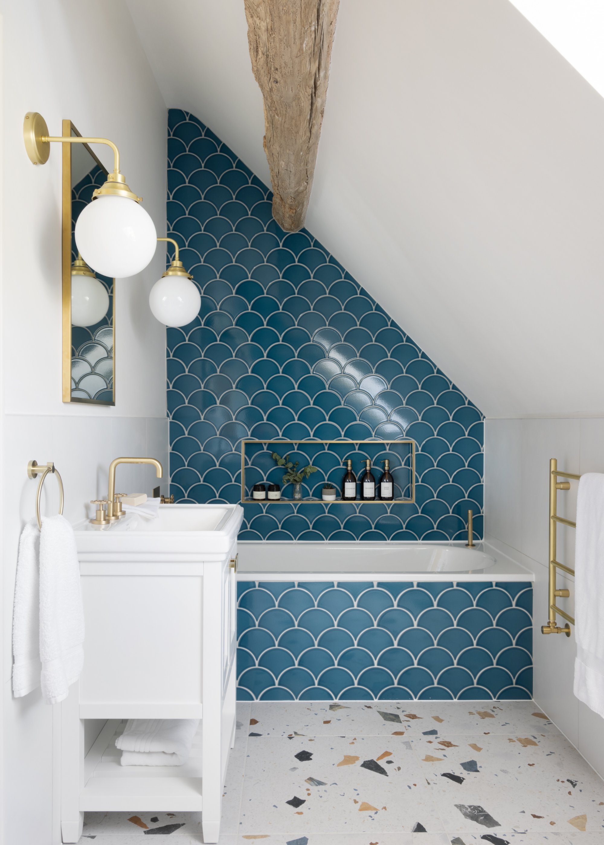 attic/ eaves bathroom with a slanted roof and a wooden beam running through. The back wall is covered in a dark blue scalloped tile and has a built-in bath tub. The floors are a multi colored terrazzo