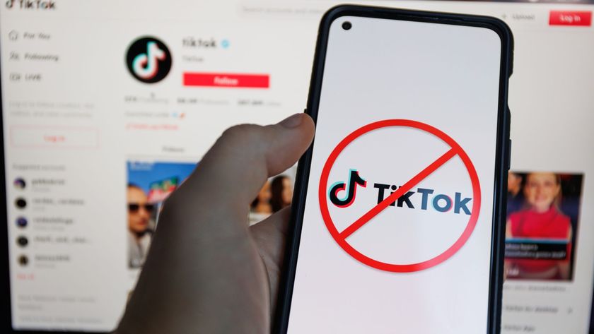 A person holding a phone with the TikTok logo crossed out displayed on the screen, with a blurry image of the tiktok homepage in the background