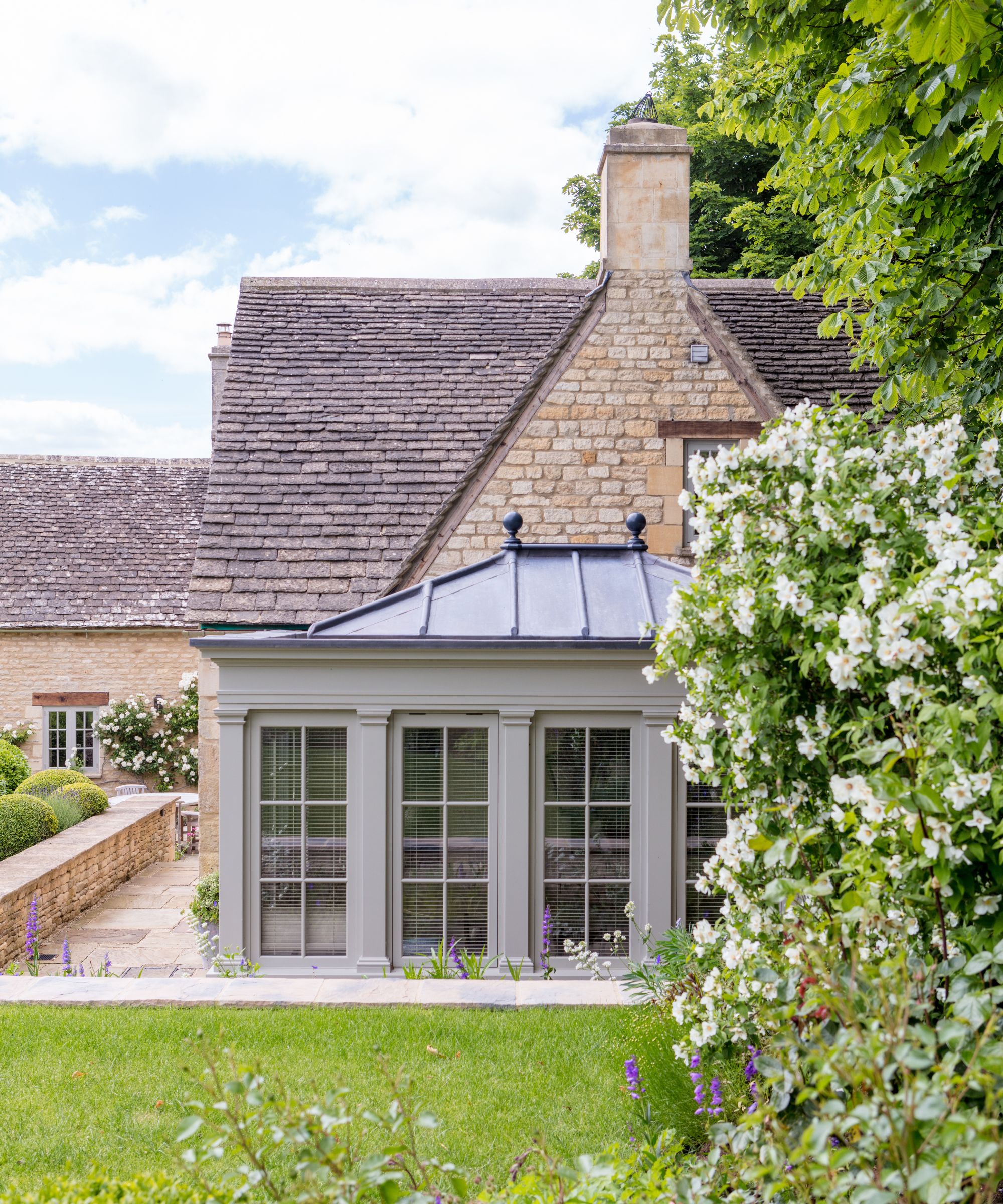 A charcoal grey orangery with a lead hip roof and black ball finials attached to a Cotswold stone cottage, framed by white climbing roses and a stone terrace.