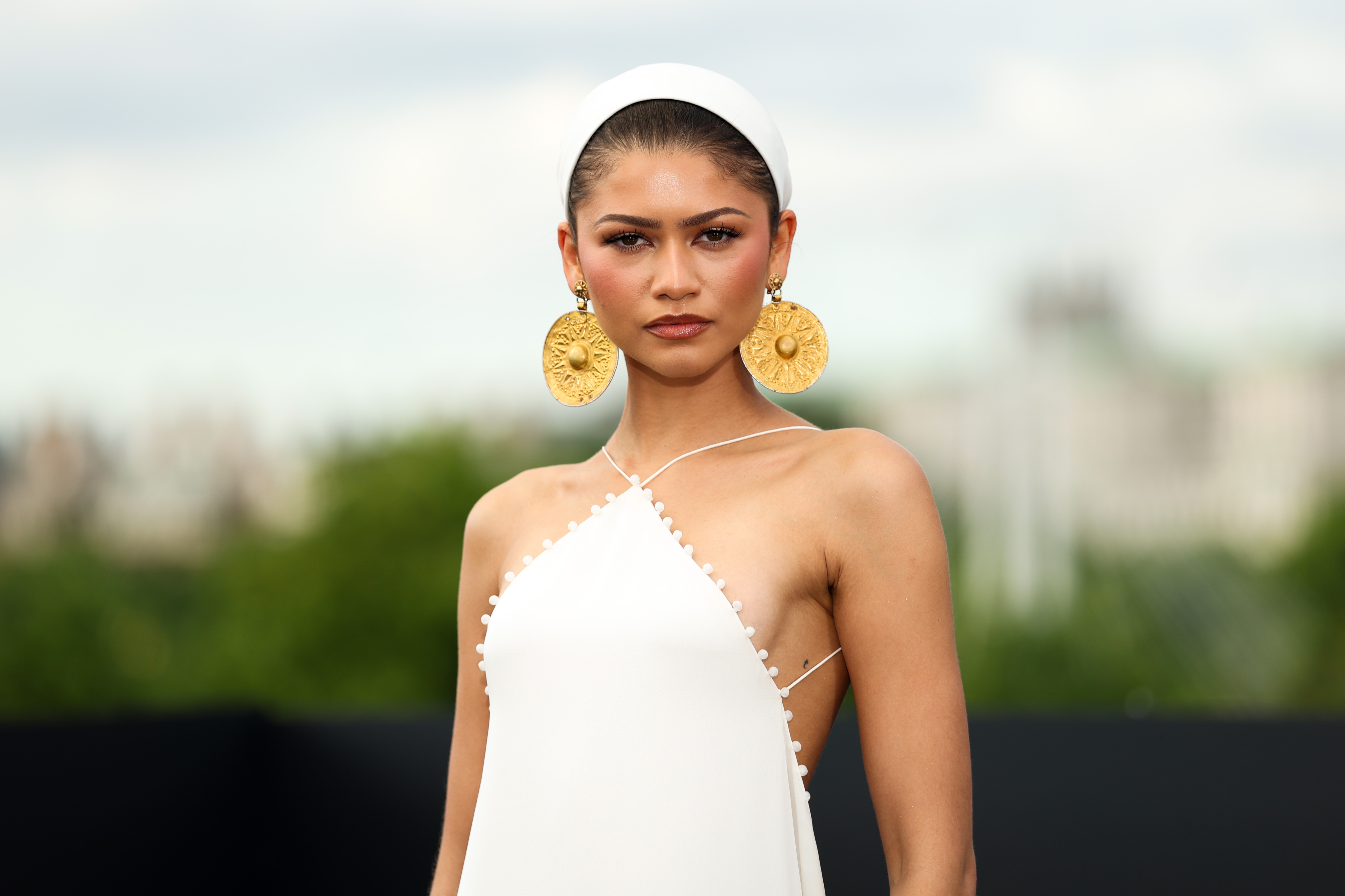 Zendaya posing on a red carpet in a white halter neck dress and large gold earrings 
