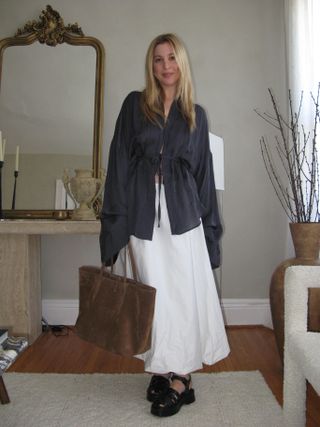 Woman standing in apartment wearing black top, white maxi skirt, brown suede tote bag, black fisherman sandals