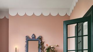 Pink wall with a scalloped white ceiling border, ornate mirror, flowers, and a dark green door frame.