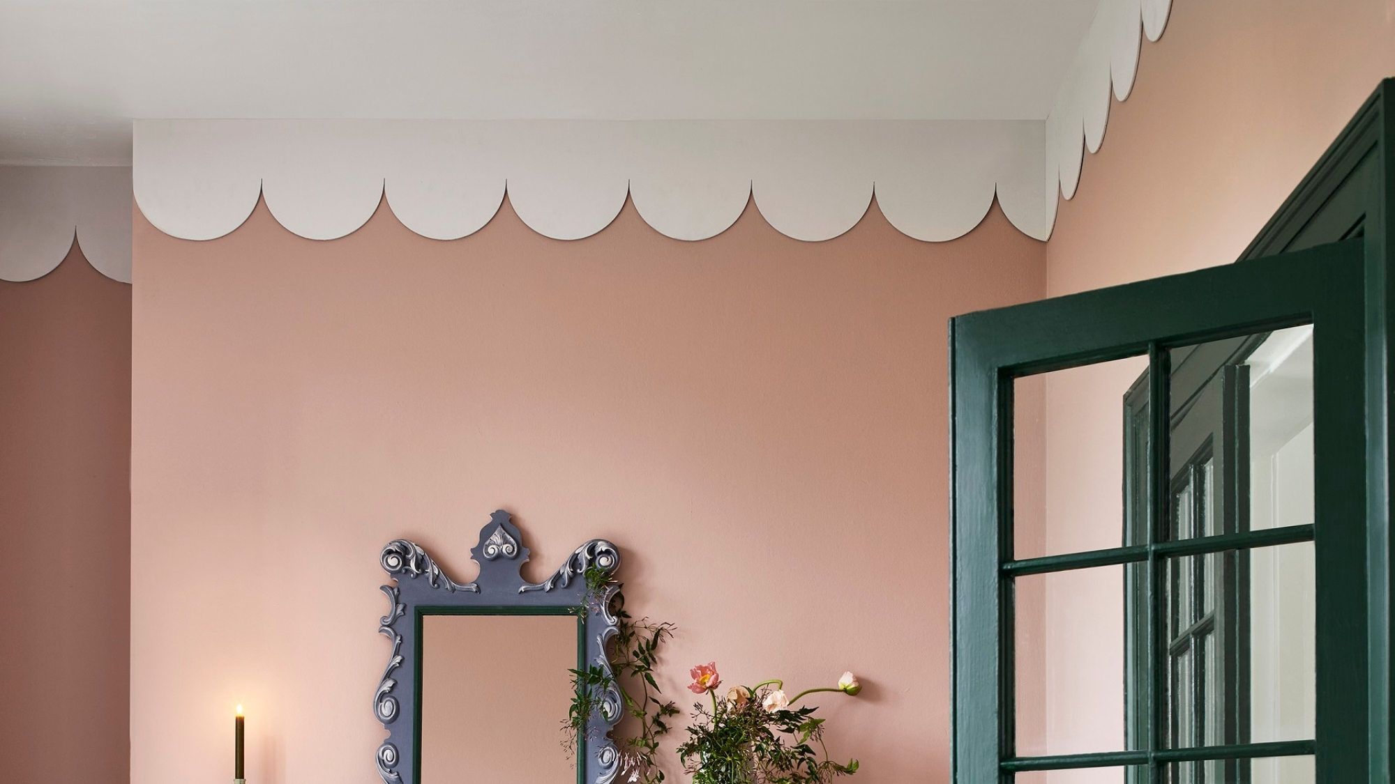 Pink wall with a scalloped white ceiling border, ornate mirror, flowers, and a dark green door frame.