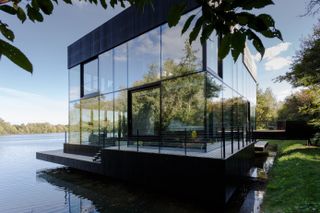 Side view of the two-floor, all-glass villa, surrounded by woods and a lake. We see a deck that goes all around the villa.