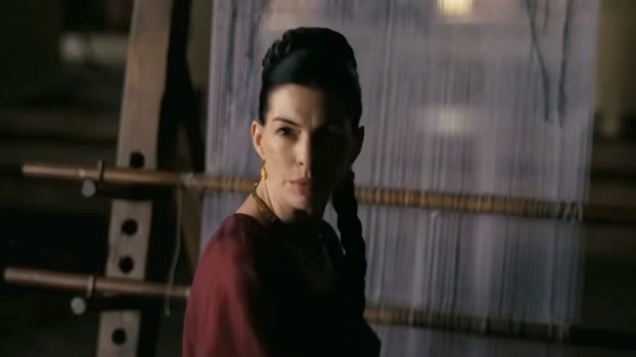 Anne Hathaway in a red dress, sitting in front of a loom in The Odyssey